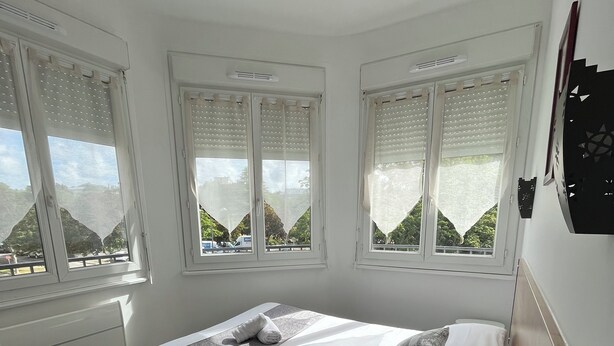 A bedroom with a bed, a bedside table with a power outlet, and large windows with blinds.