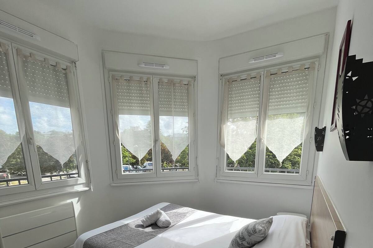 A bedroom with a bed, a bedside table with a power outlet, and large windows with blinds.