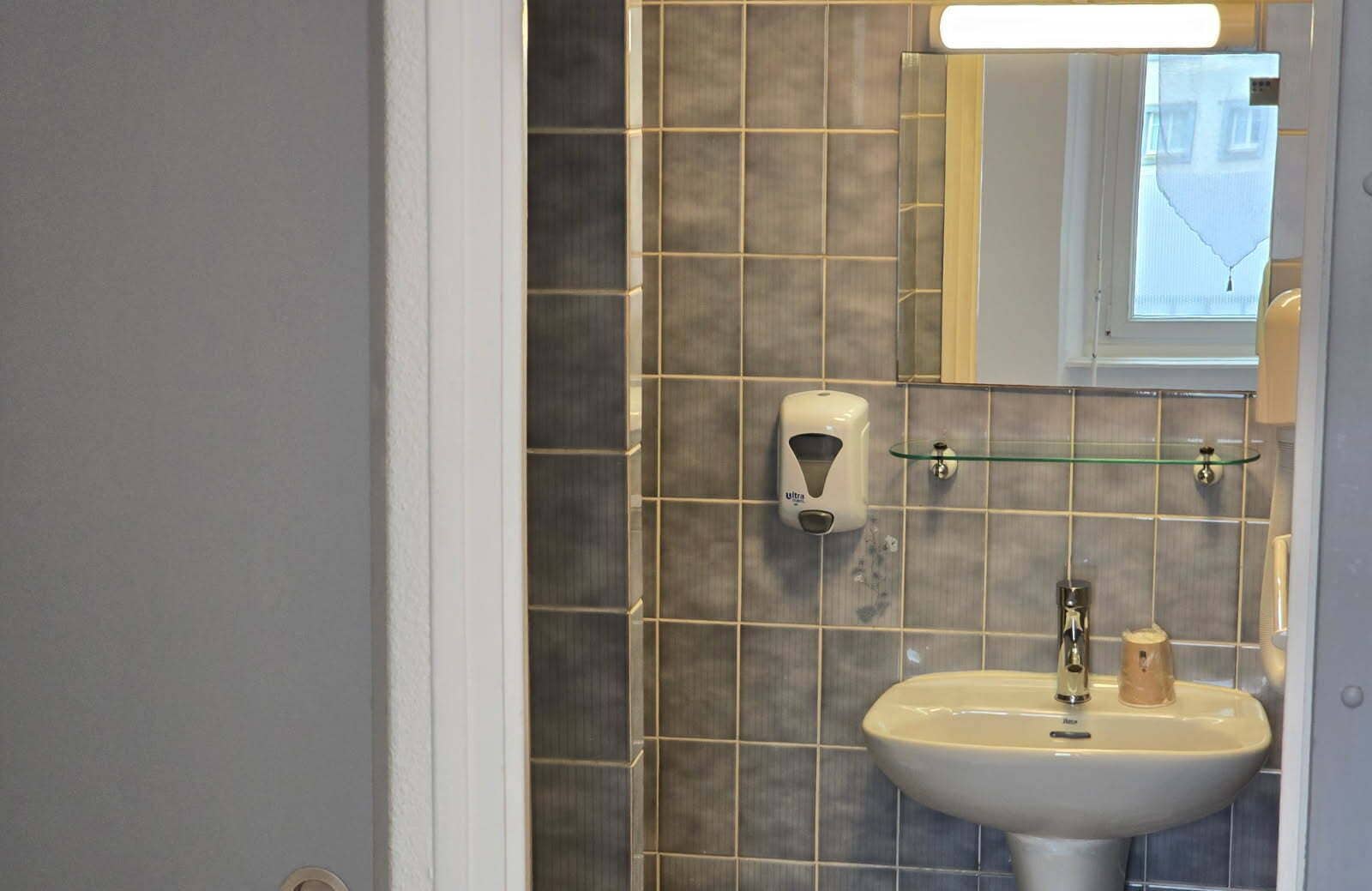 A bathroom with a white sink, a soap dispenser, and a glass shelf.