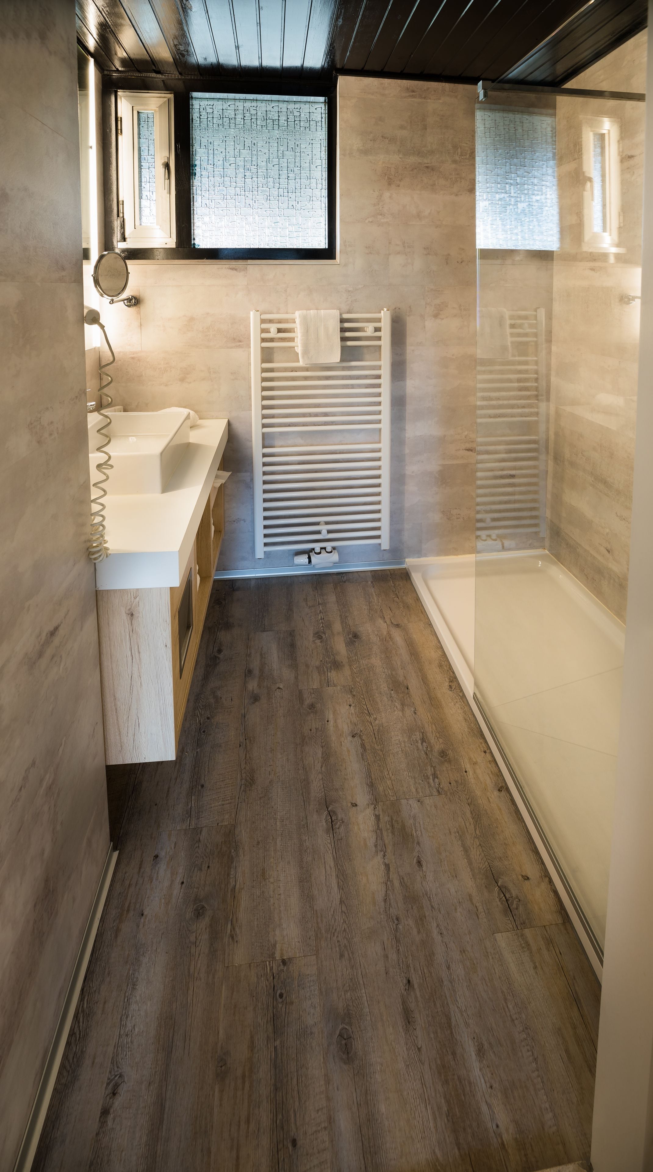 A modern bathroom with a wooden floor, a white sink, a towel rack, and a glass shower enclosure.