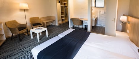 A hotel room with a large bed, two armchairs, a small table, a desk, a wardrobe, and a bathroom with a mirror and sink.