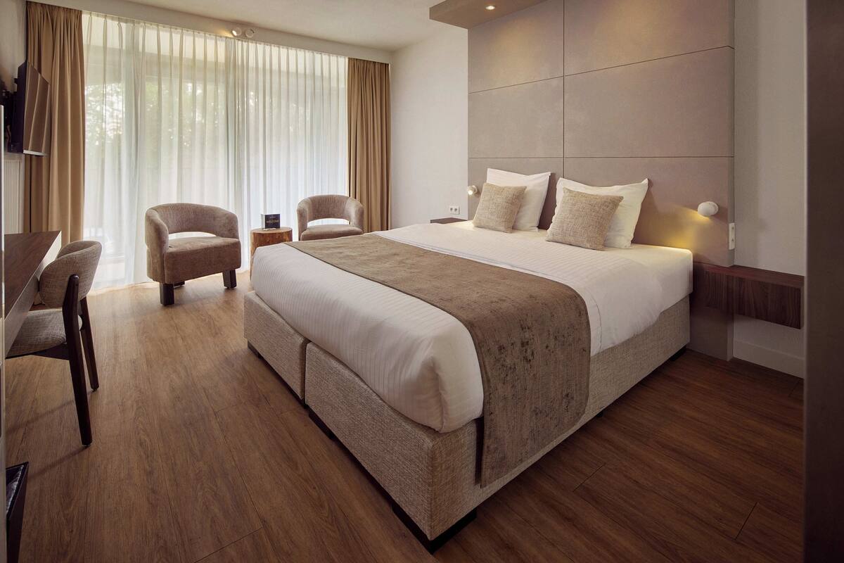 A modern hotel room with a large bed, two armchairs, a desk, and a large window with curtains.
