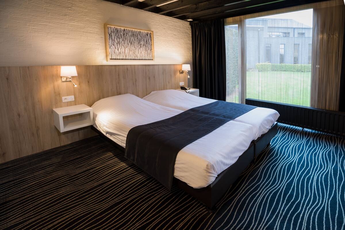 A hotel room with a large bed, two bedside tables, a wall-mounted lamp, and a view of the outside.