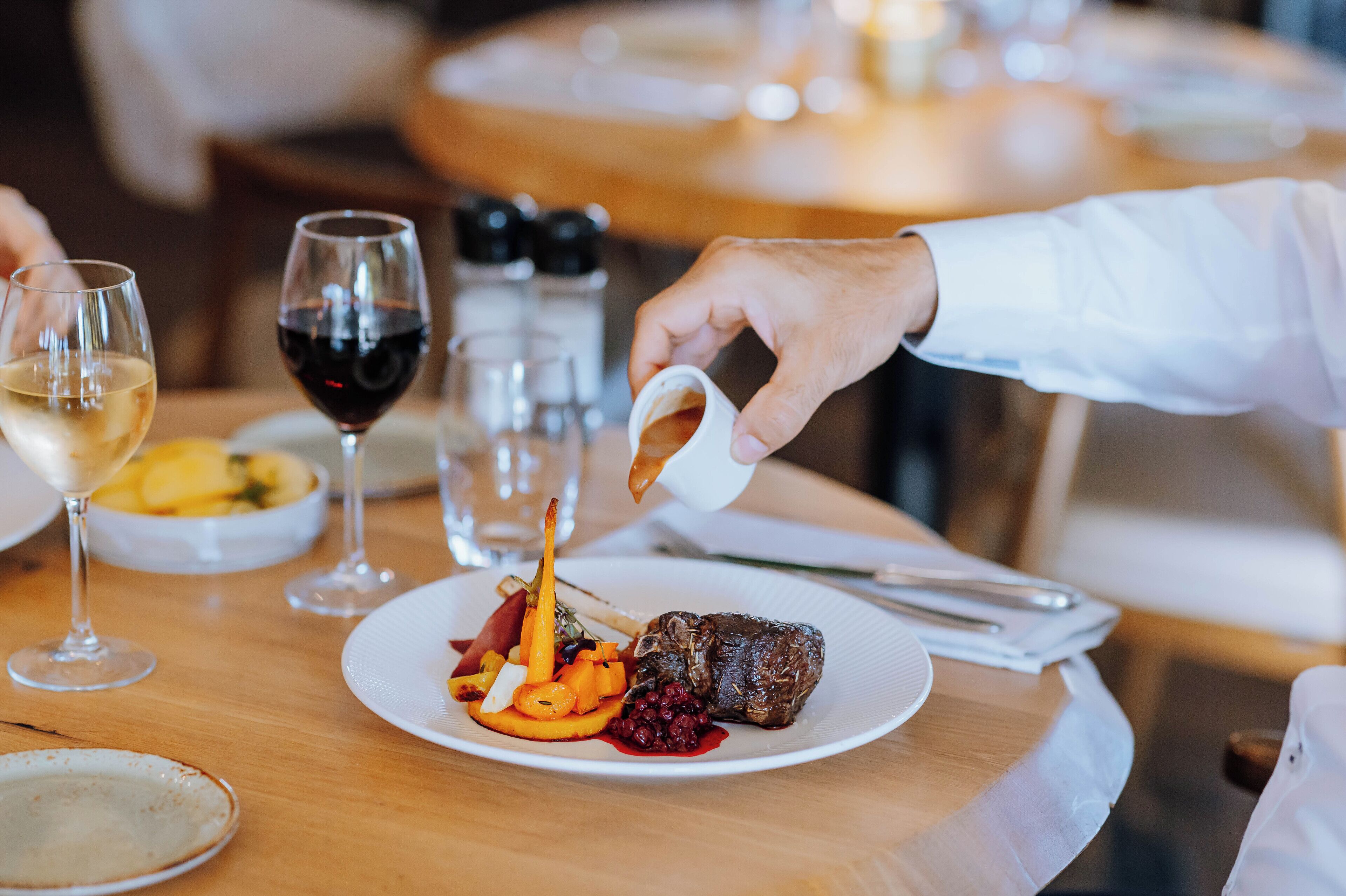 A dining table with a plate of food, a glass of red wine, and a hand pouring sauce.