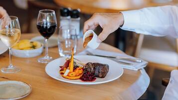 A dining table with a plate of food, a glass of red wine, and a hand pouring sauce.