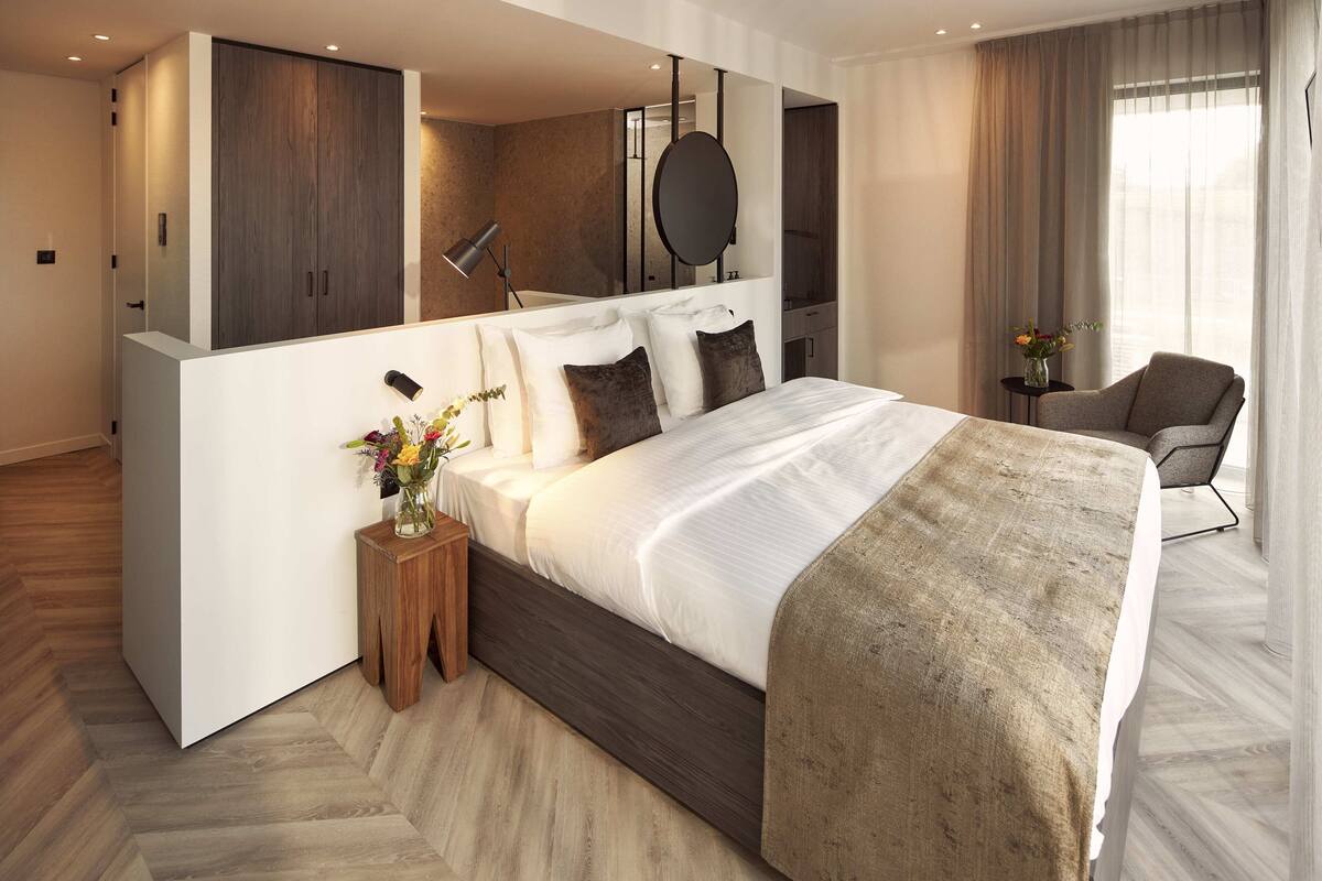 A modern hotel room with a large bed, a side table with a vase of flowers, a chair, and a large mirror above the bed.
