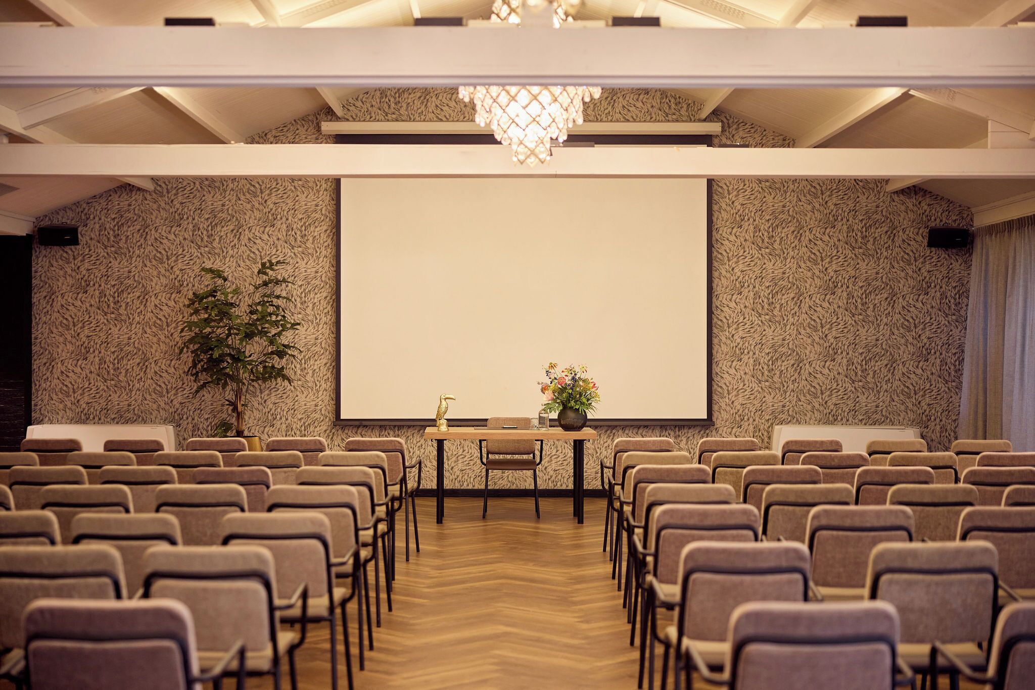 A conference room with rows of chairs, a projector screen, a table with a vase of flowers, and a plant.