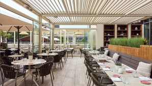 An outdoor dining area with round tables and chairs, a wooden bench, and a glass wall. - Van Der Valk Hotel Beveren (Beveren)