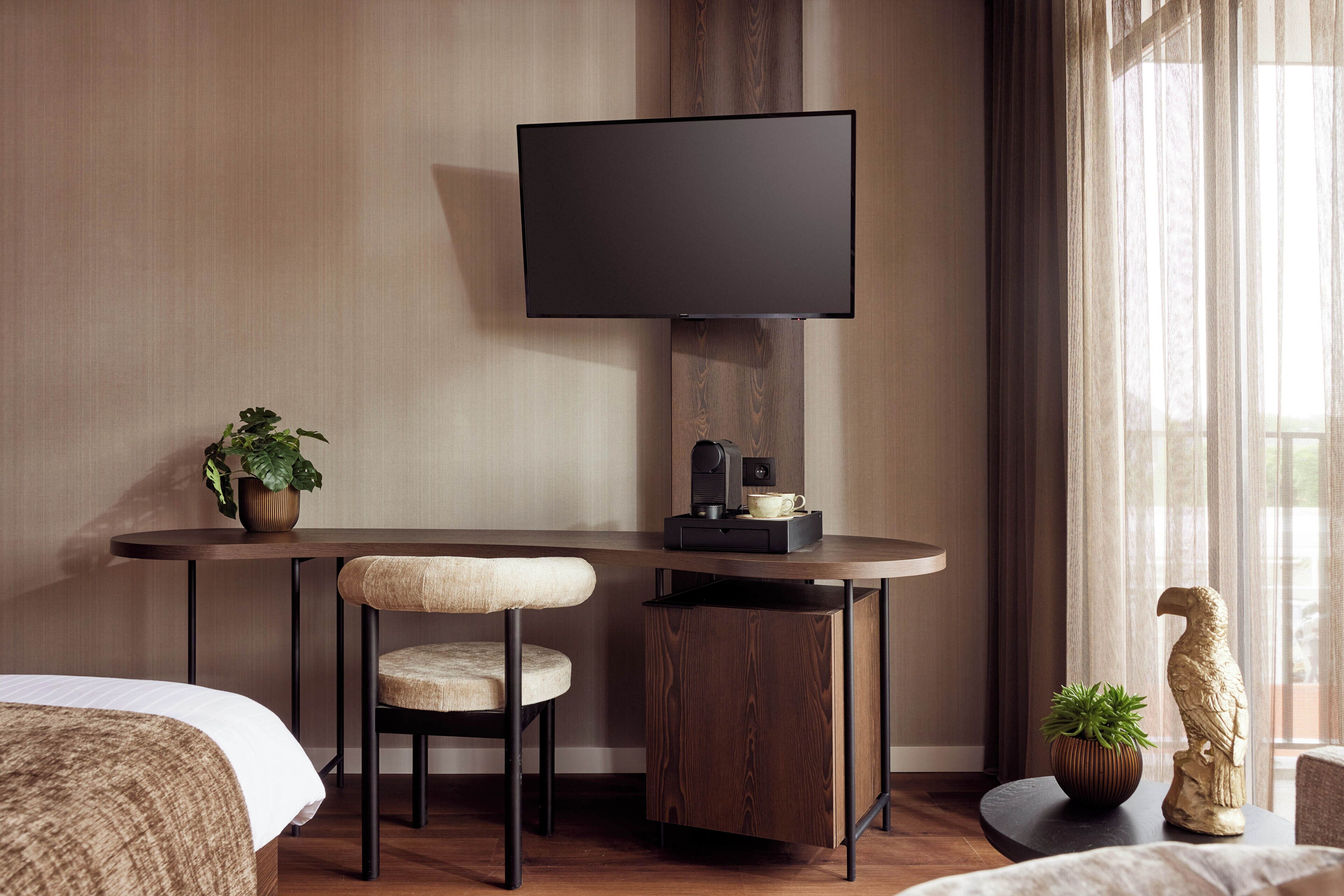 Habitación de hotel con cama, escritorio, silla, televisor, cafetera y planta en maceta.