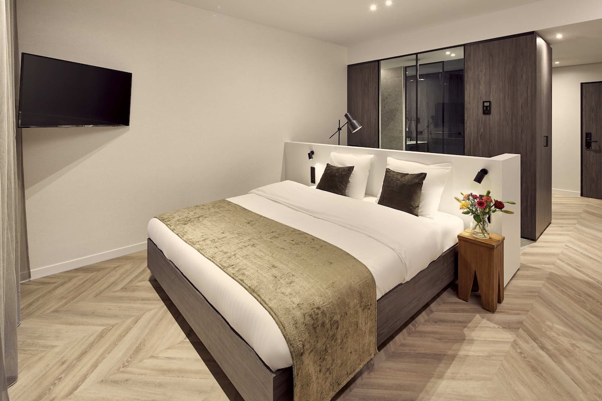 A modern bedroom with a large bed, a TV mounted on the wall, a bedside table with a vase of flowers, and a wardrobe with a mirror.
