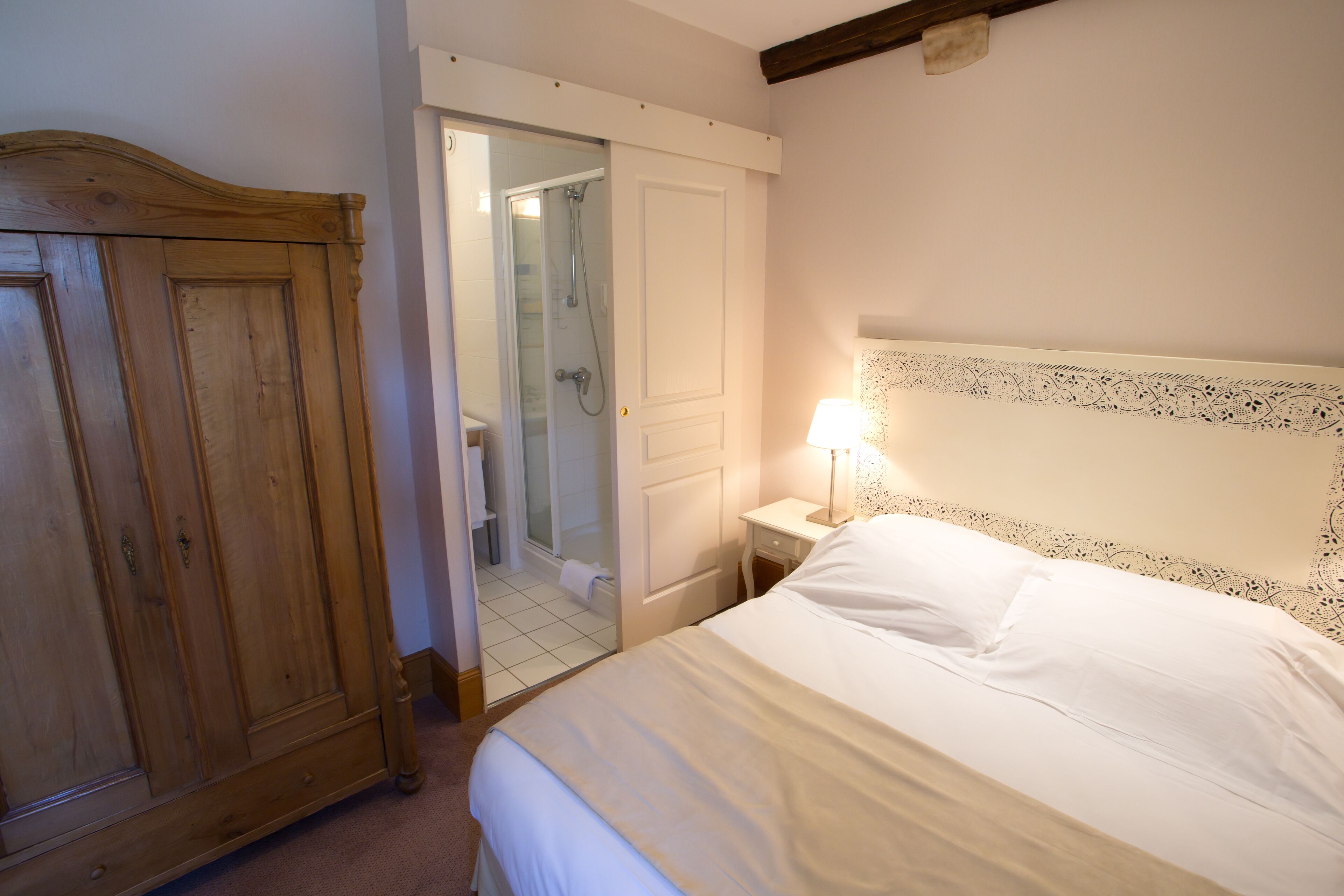 A bedroom with a wooden wardrobe, a bedside table with a lamp, and a bathroom visible through an open door.