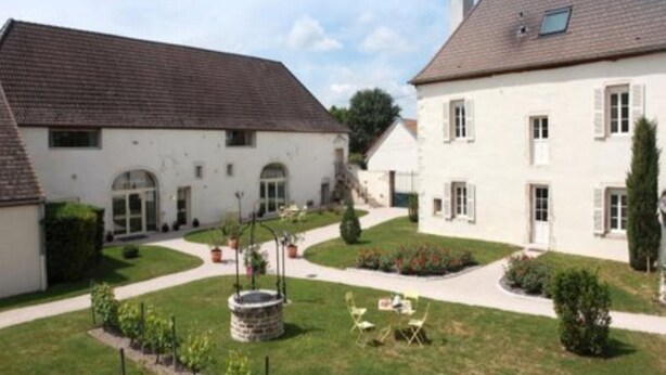 Un patio con dos edificios blancos, una fuente de piedra y un pequeño jardín con sillas.