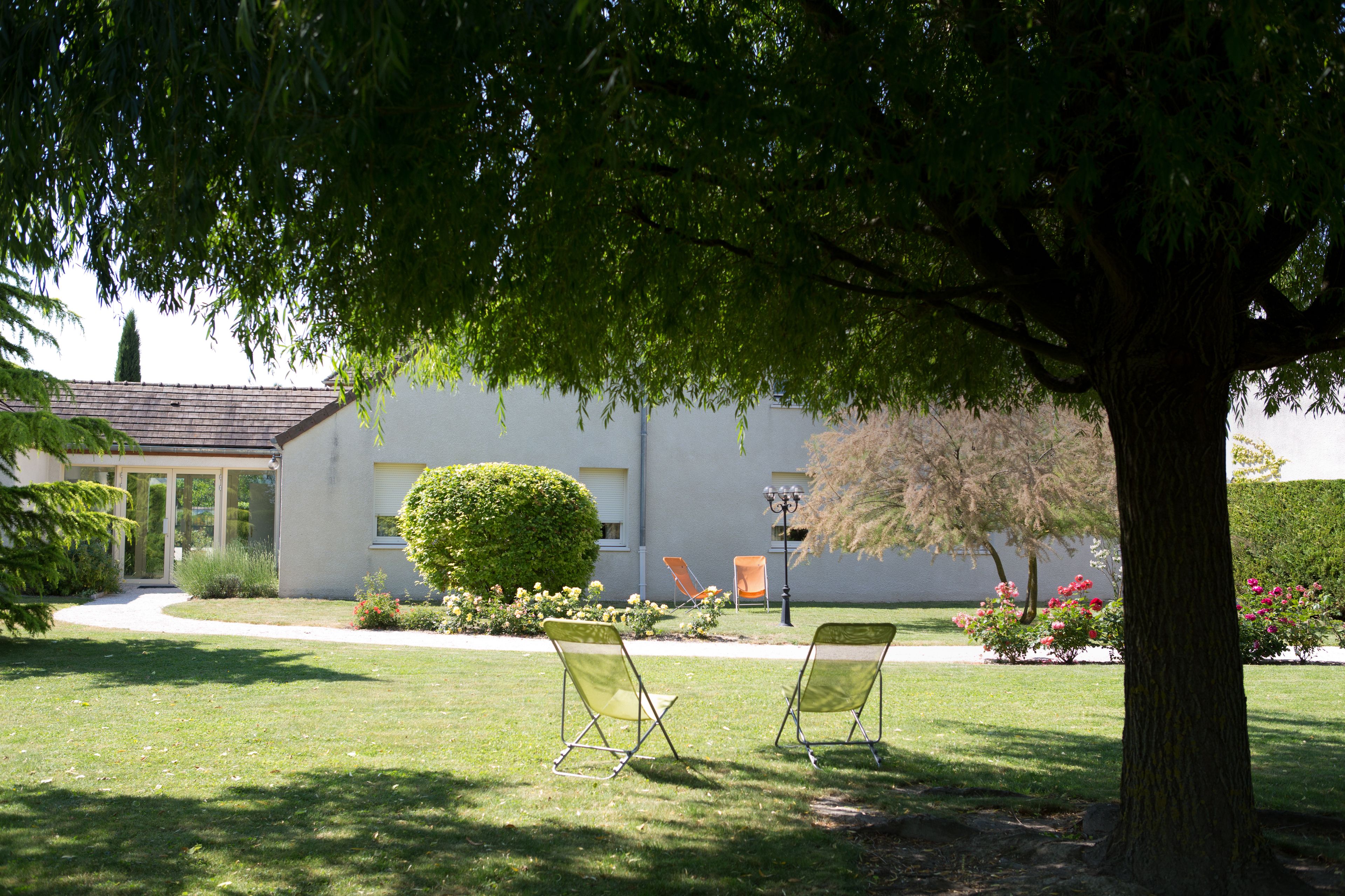 A building with a white exterior, a paved driveway, and a well-maintained lawn. There are two lawn chairs under a tree.