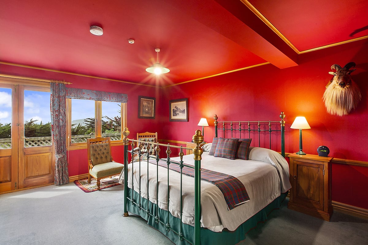 A bedroom with a four-poster bed, a wooden chair, a nightstand with a lamp, and a mounted animal head on the wall.