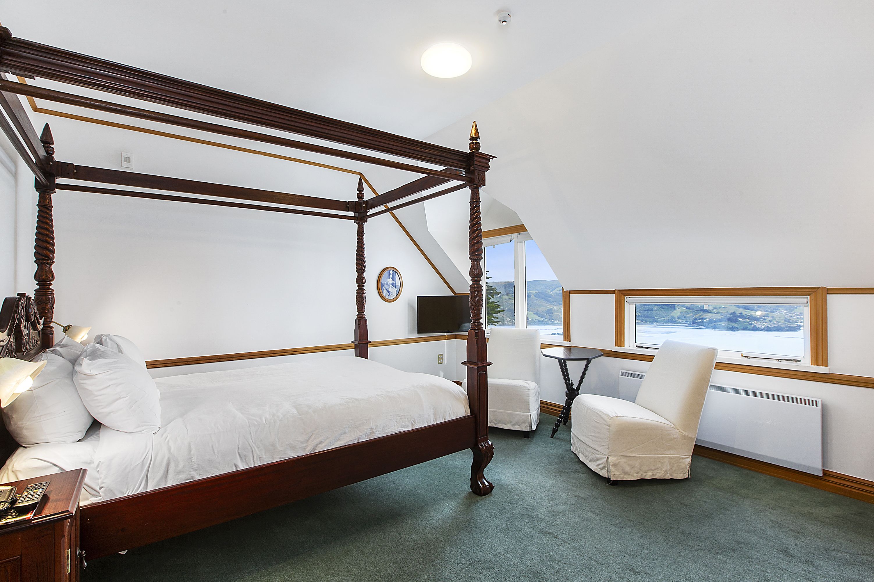 A four-poster bed with a wooden frame, a bedside table with a lamp, a chair, and a view of the outdoors through a window.