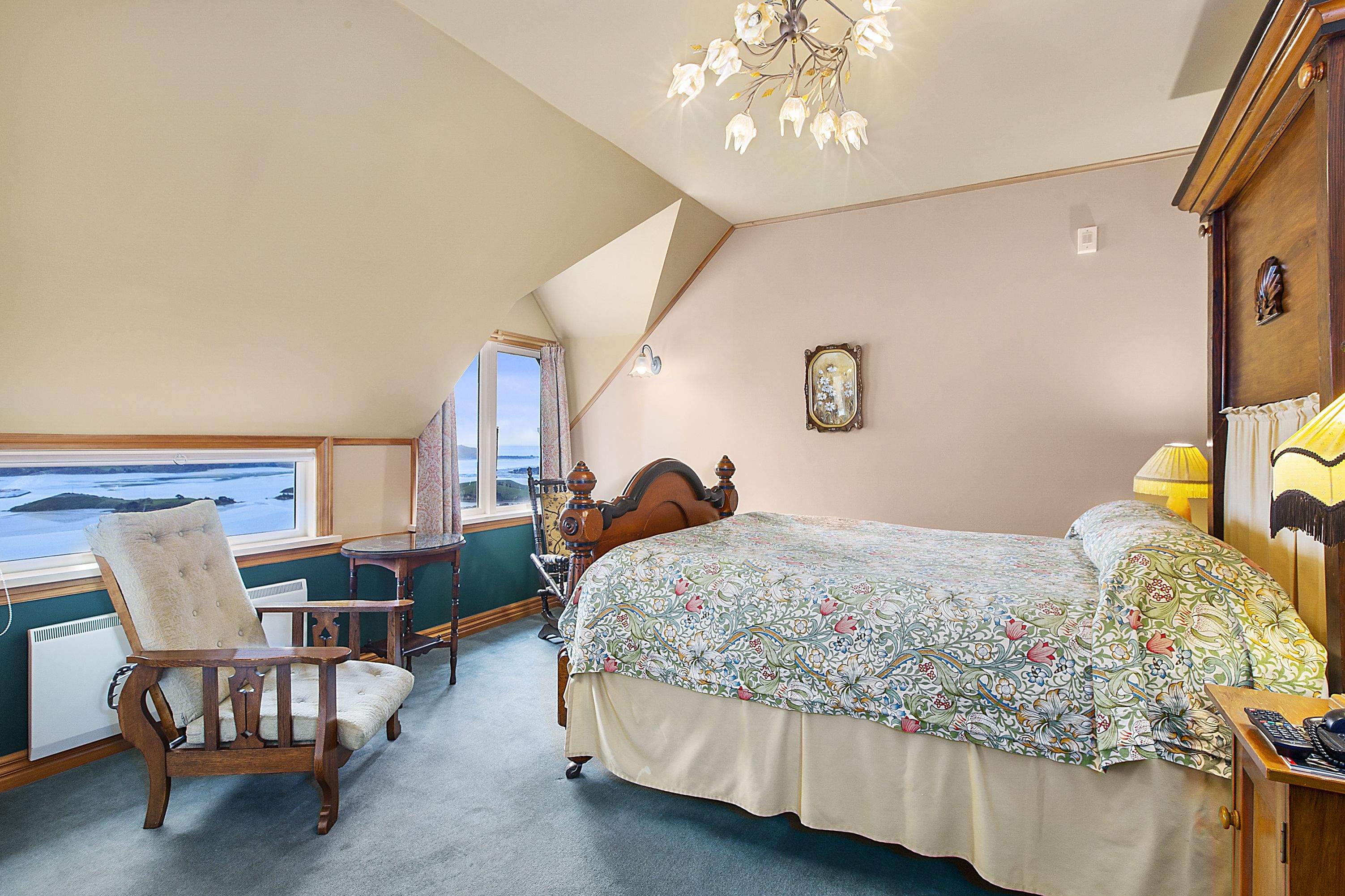 A bedroom with a large bed, a chair, a small table, a lamp, and a view of the sea through a window.