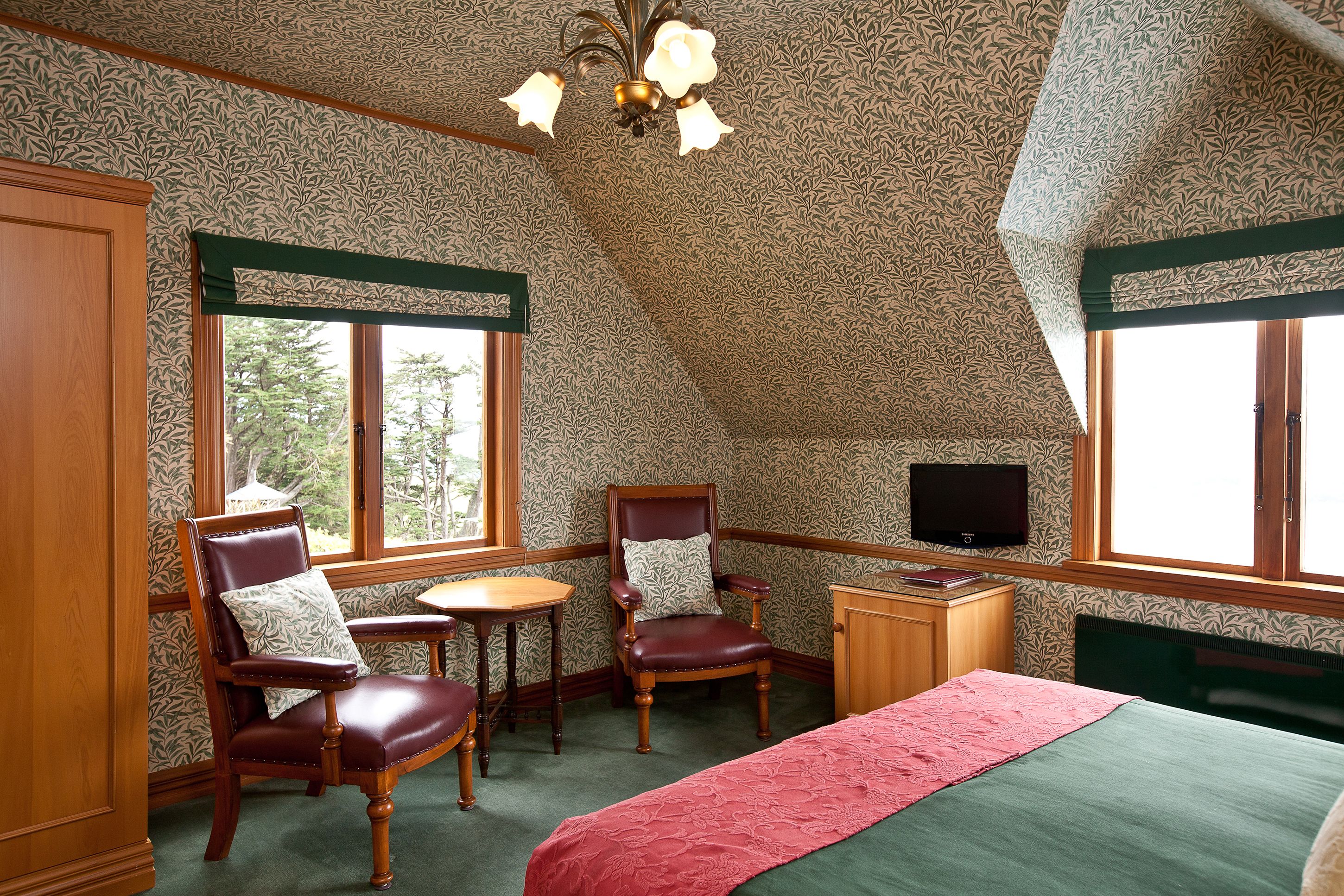 A bedroom with a large bed, two chairs, a small table, a television, and large windows.