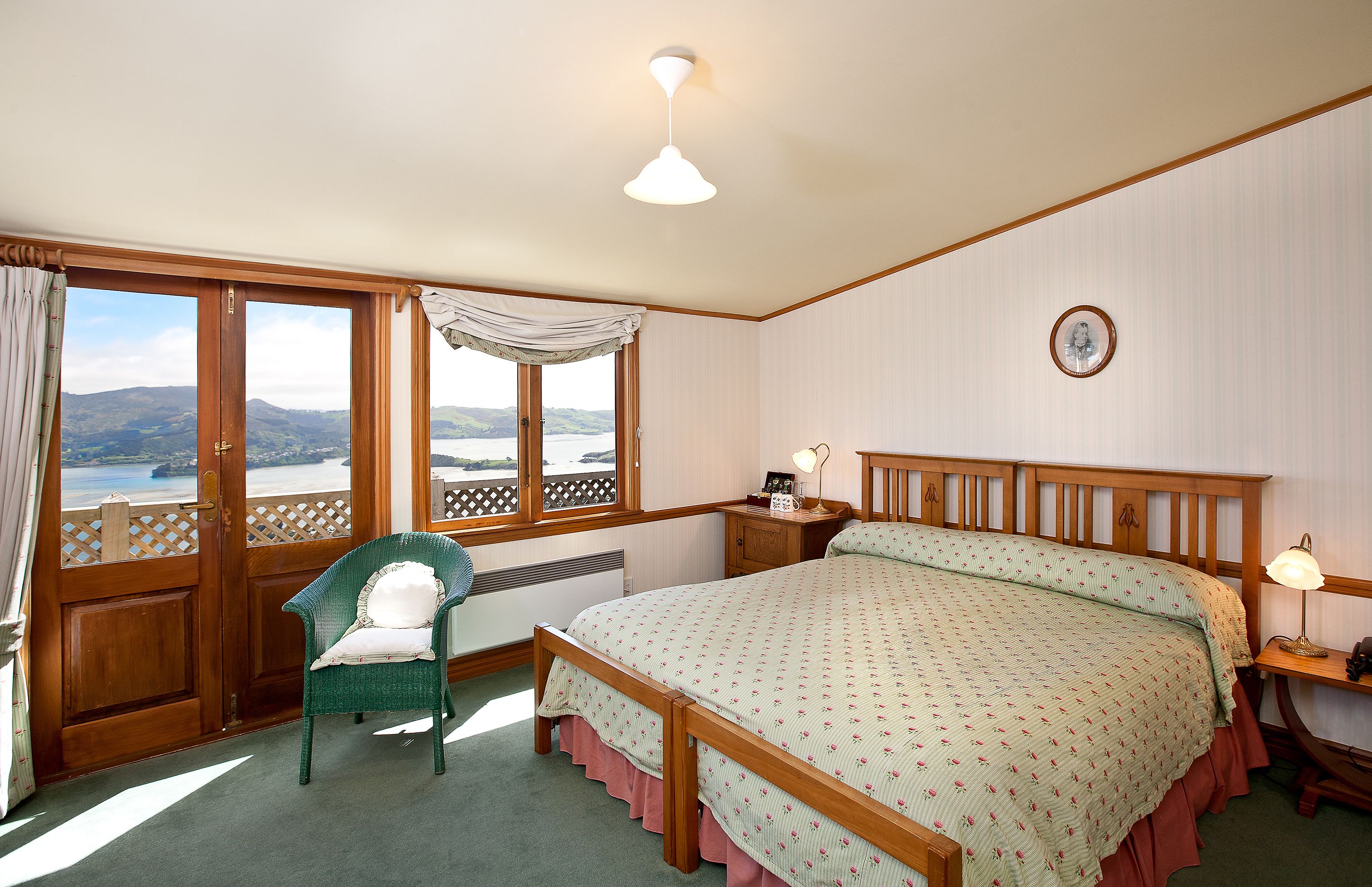 A bedroom with a large bed, a chair, a nightstand, a lamp, and a view of the outdoors.