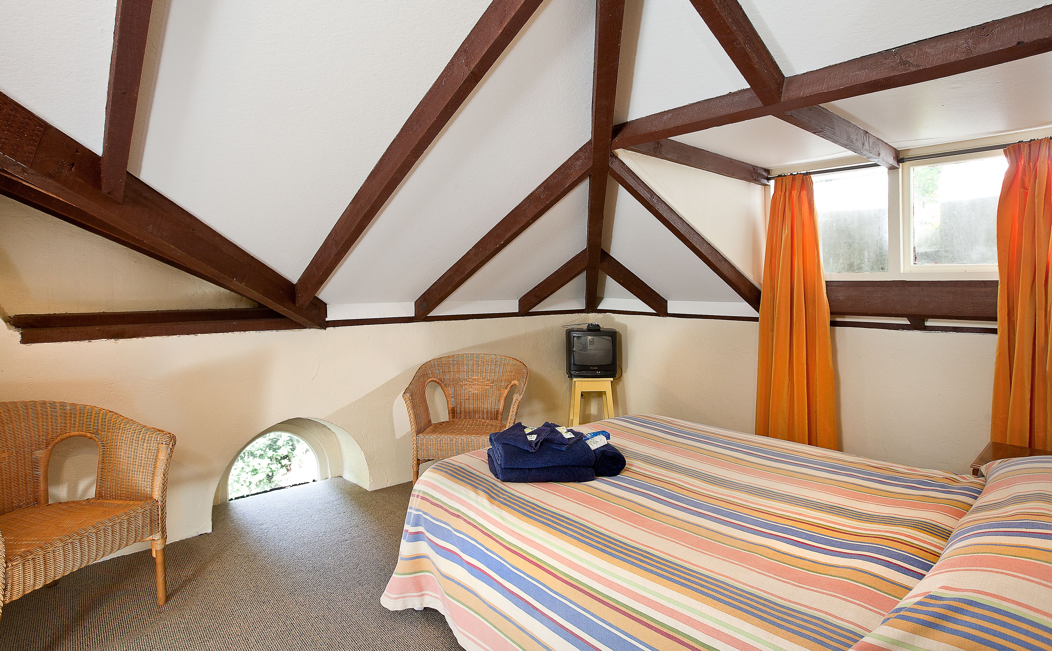 Un dormitorio con techo inclinado, una cama con colcha a rayas, una silla de mimbre, una mesita con un televisor y una ventana con cortinas.