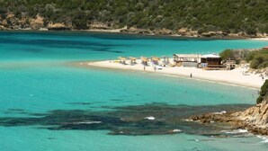 A coastal beach with clear turquoise waters, a sandy shore, and a small building with umbrellas and a lifeguard stand. - Gentarrubia - B&B (Capoterra)