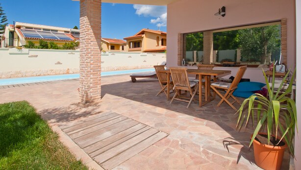 A covered patio with a stone floor, a dining table with chairs, a large window, and a potted plant. - Gentarrubia - B&B (Capoterra)