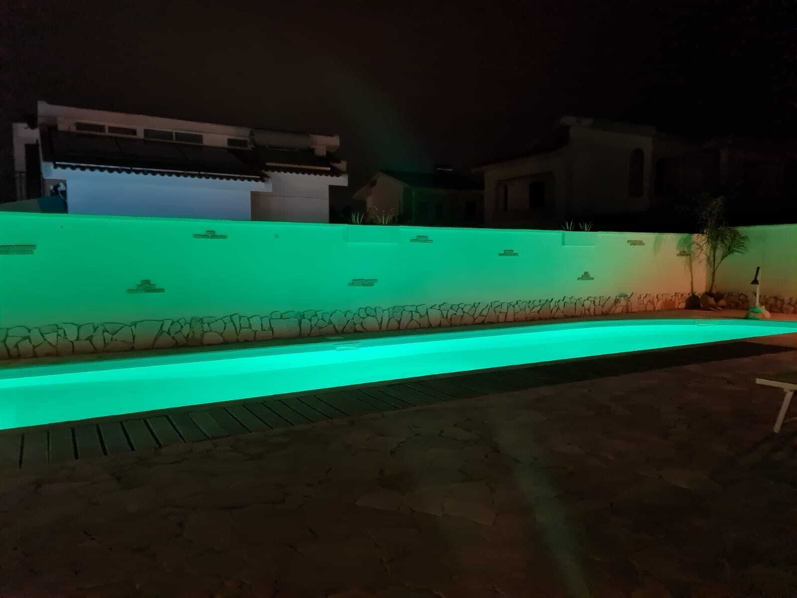Una piscina con un borde iluminado en verde, rodeada por un muro de piedra y una zona pavimentada.