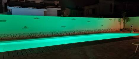 A swimming pool with a green-lit edge, surrounded by a stone wall and a paved area.