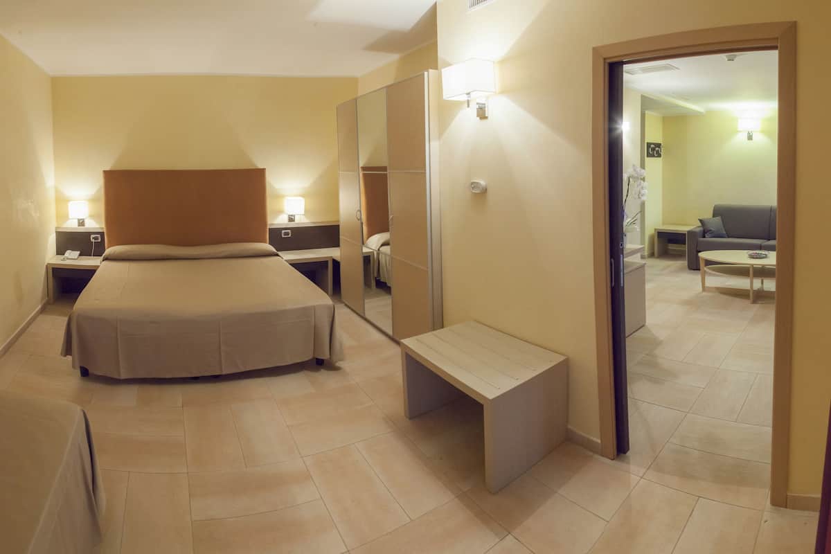 Habitación de hotel con cama, mesitas de noche, un banco, un espejo y vistas al área de estar contigua.
