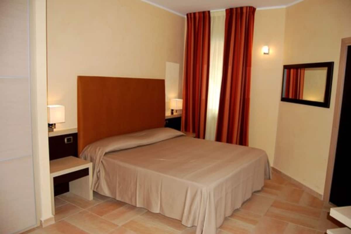 Habitación de hotel con una cama individual, una mesita de noche, una lámpara, un espejo y una ventana con cortinas.