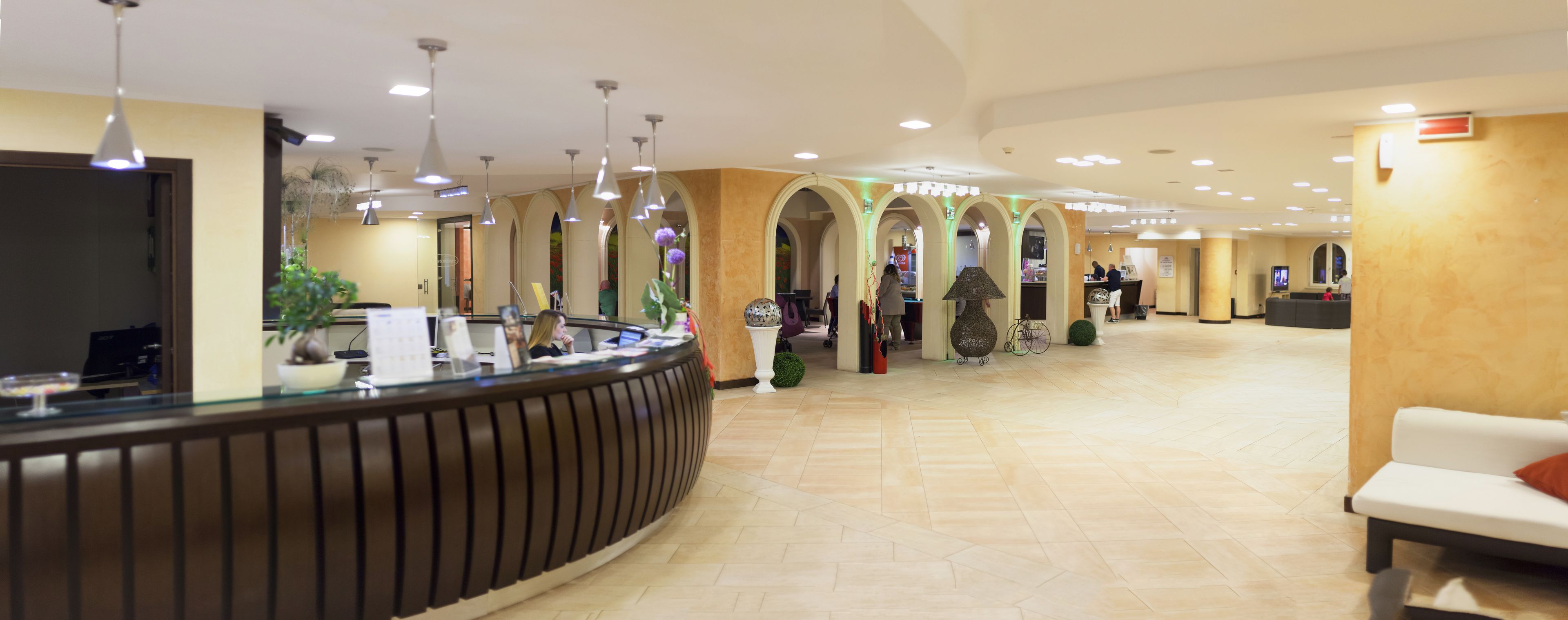 Un'area reception di un hotel con una scrivania a reception a forma di arco, diverse luci a sospensione e un'area salotto con un divano bianco.
