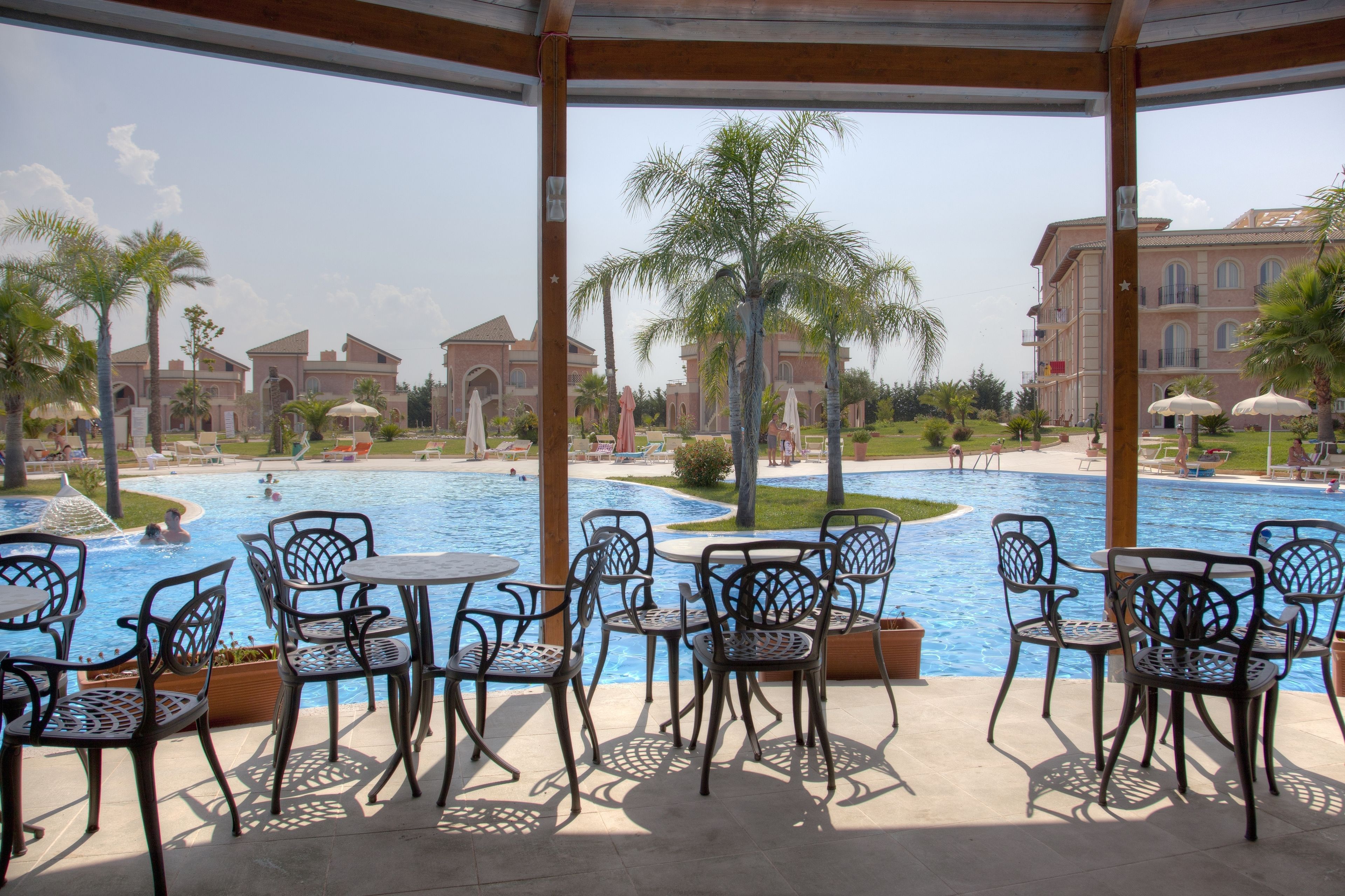 Zona junto a la piscina con sillas y mesas, una piscina y vistas a un edificio con palmeras.