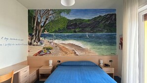 A bedroom with a large mural of a beach scene, a bed with a blue bedspread, a desk, and a chair. - Hotel Aquavite (Gardone Riviera)