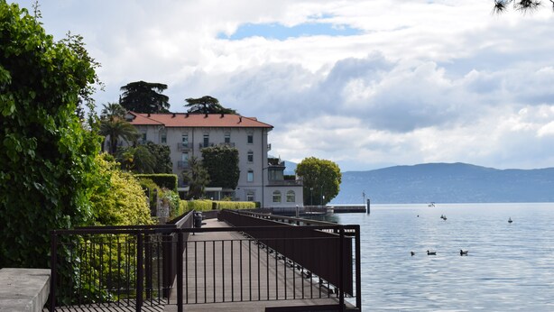 A waterfront property with a wooden dock, a large building with a red roof, and a calm lake with boats. - Hotel Aquavite (Gardone Riviera)