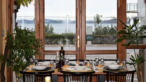 A dining area with tables set for four, large windows offering a view of a body of water and mountains, and potted plants on either side of the tables. - Hotel Aquavite (Gardone Riviera)