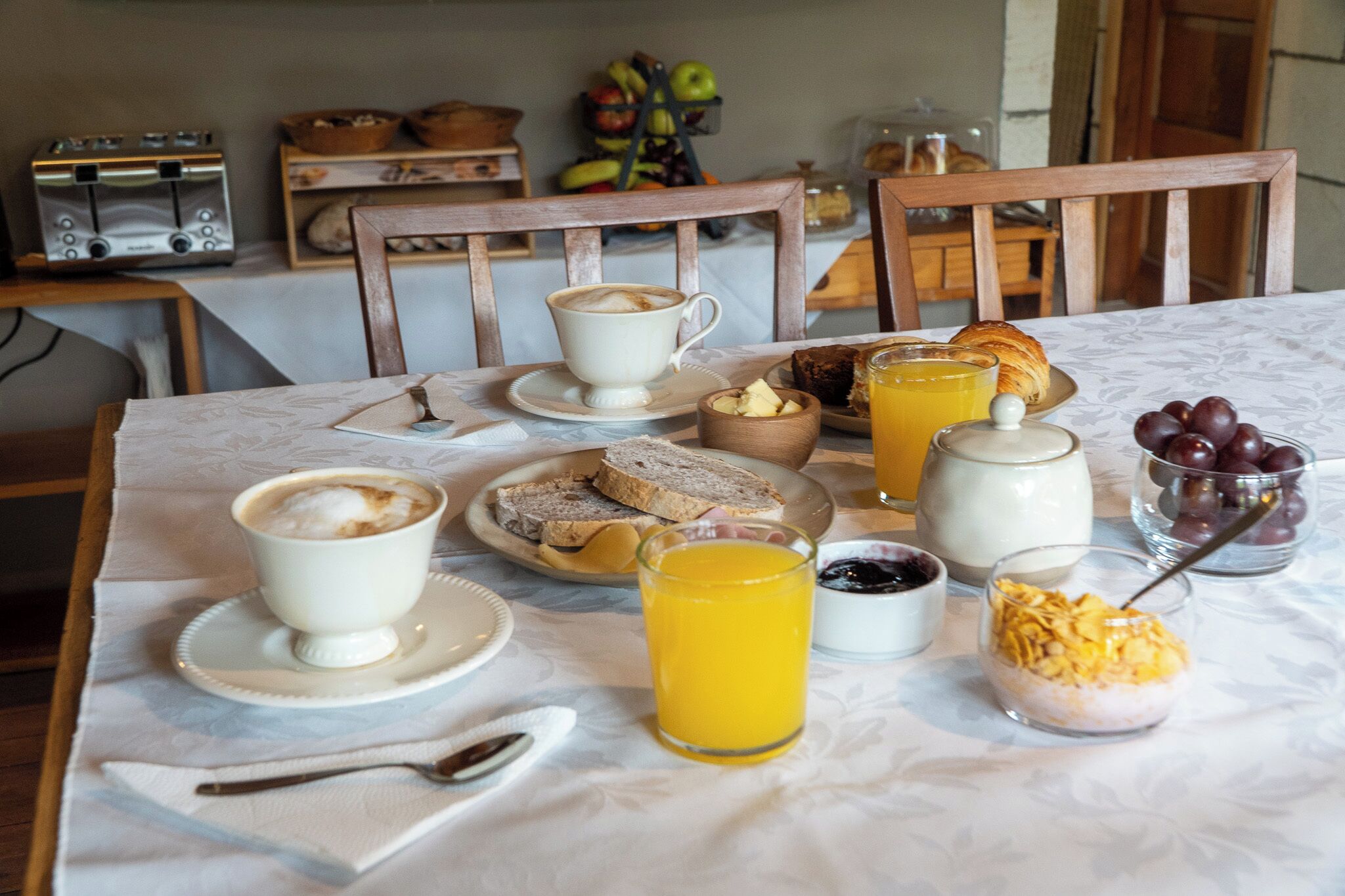 A breakfast table with croissants, a cup of coffee, orange juice, and a bowl of fruit.