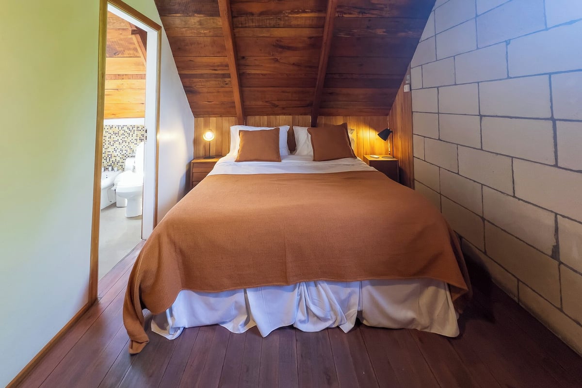 A bedroom with a wooden ceiling, a large bed with a brown bedspread, two bedside tables with lamps, and a view of a balcony.