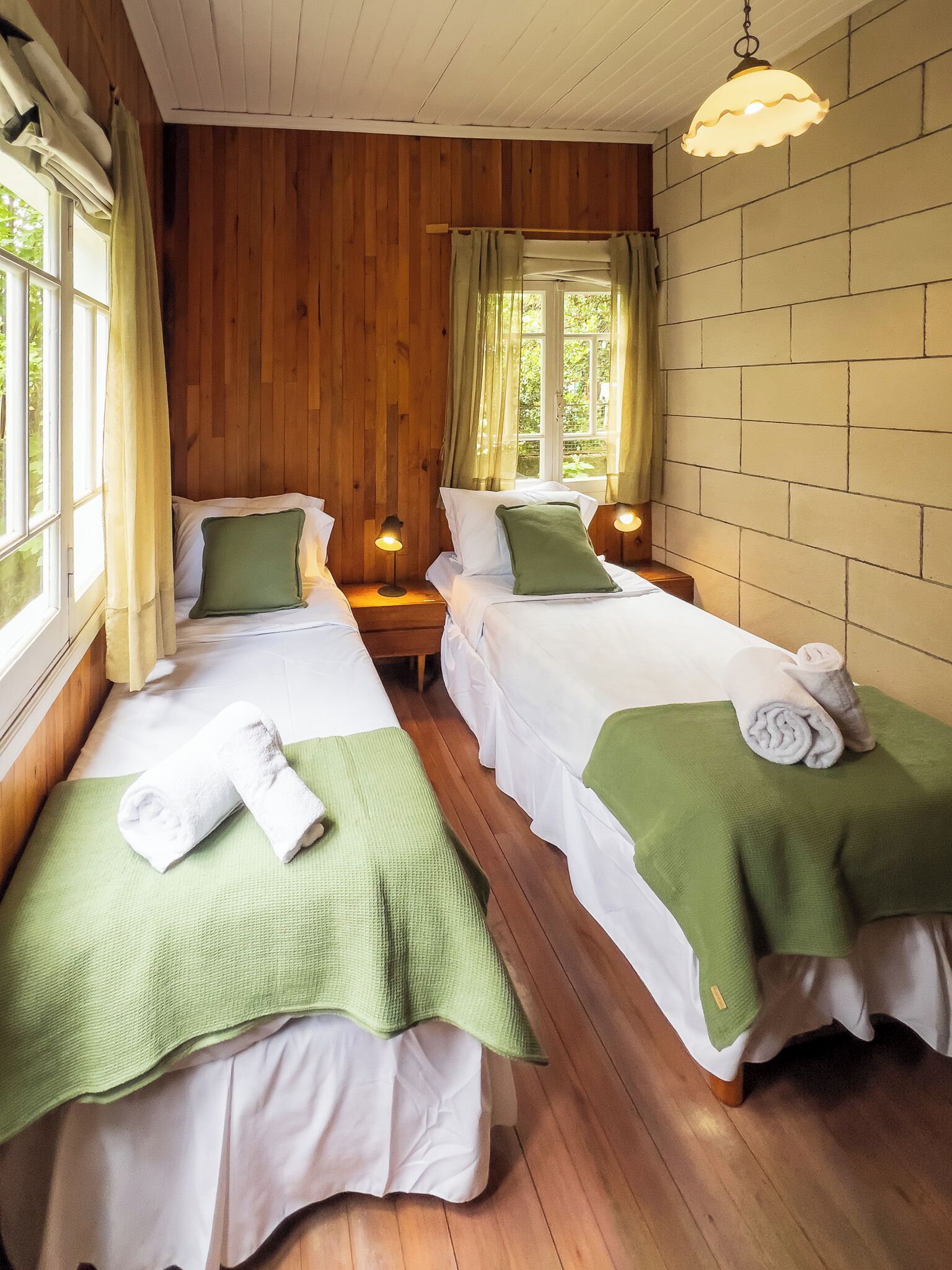 Une chambre avec deux lits simples, des murs en bois, une fenêtre avec des rideaux et un luminaire suspendu.