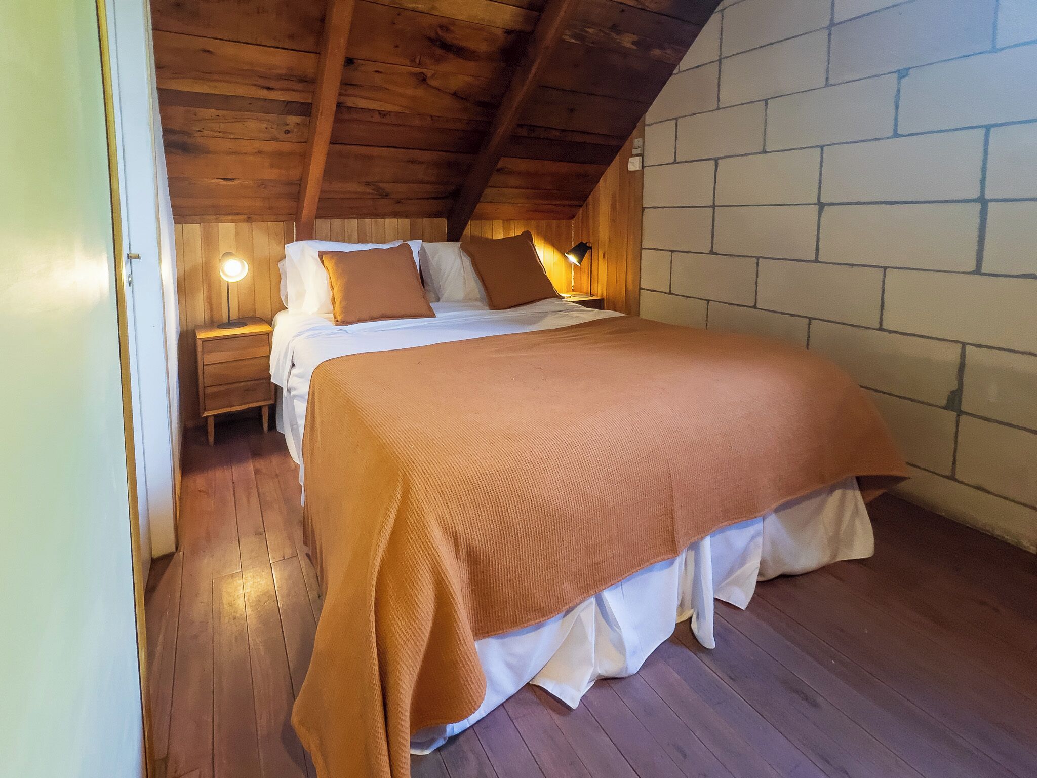 A cozy bedroom with a wooden ceiling, a brick wall, and a bed with a brown blanket.