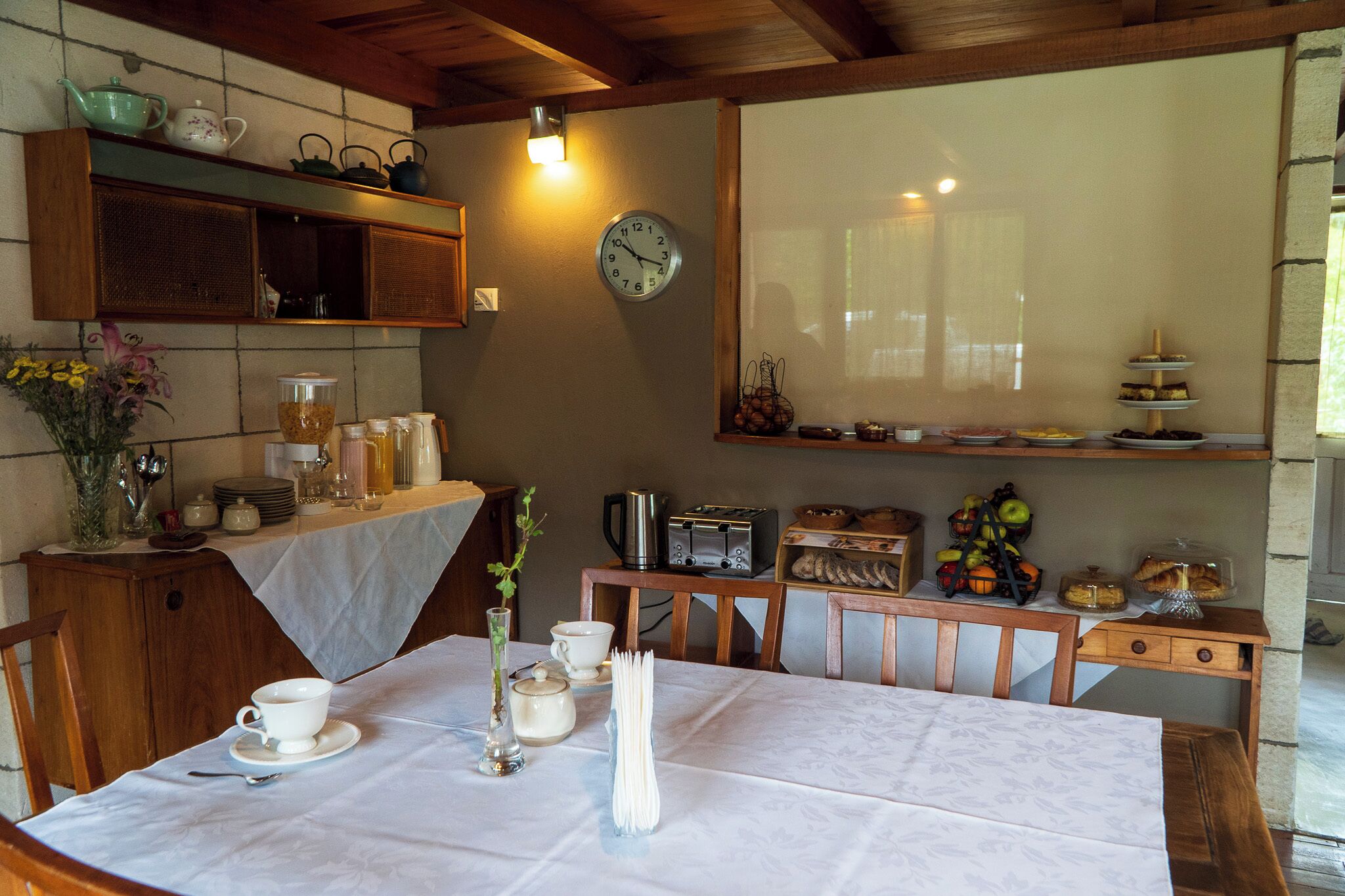 Un comedor con una mesa dispuesta para dos personas, un reloj en la pared y varios electrodomésticos y utensilios de cocina sobre la encimera.