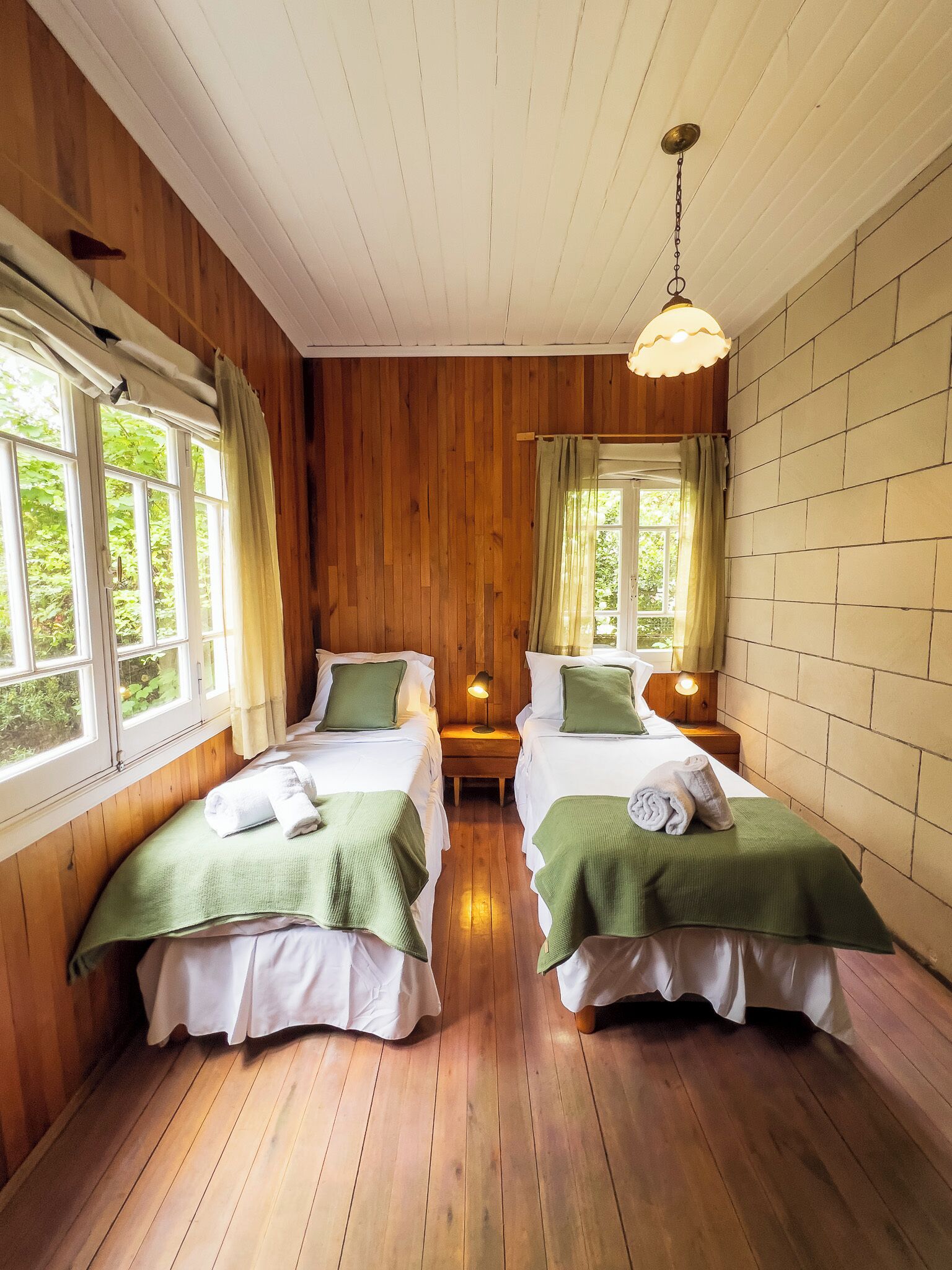 Una habitación con dos camas, paredes de madera, una lámpara colgante y ventanas con cortinas.