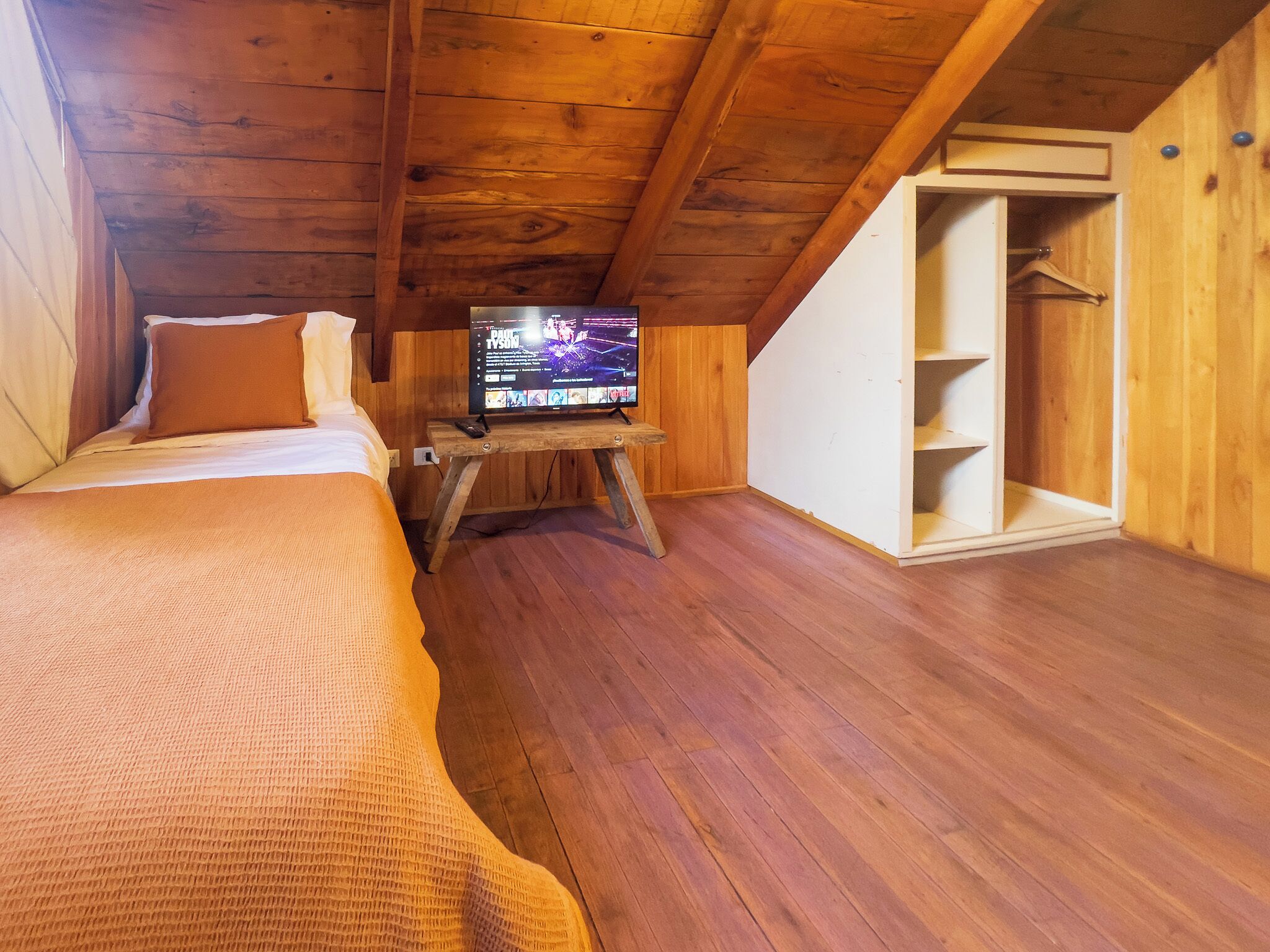 Une chambre mansardée chaleureuse avec un plancher en bois, un lit recouvert d’une couverture marron, un téléviseur posé sur un meuble en bois et une armoire.