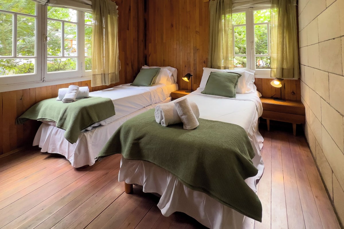 A room with two single beds, wooden floors, and windows with curtains.