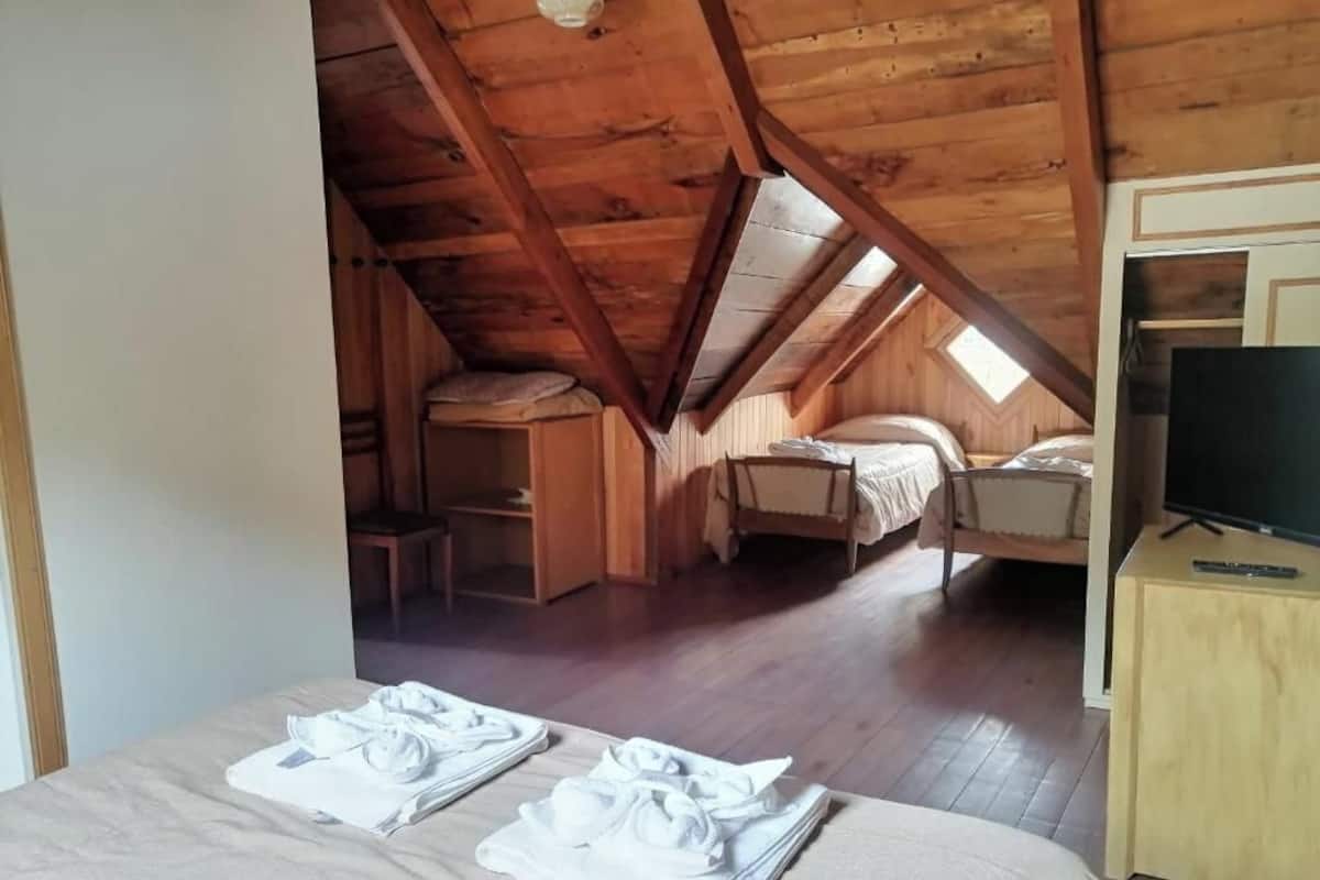 A cozy attic bedroom with a wooden ceiling, two beds, a desk, and a TV on a wooden cabinet.