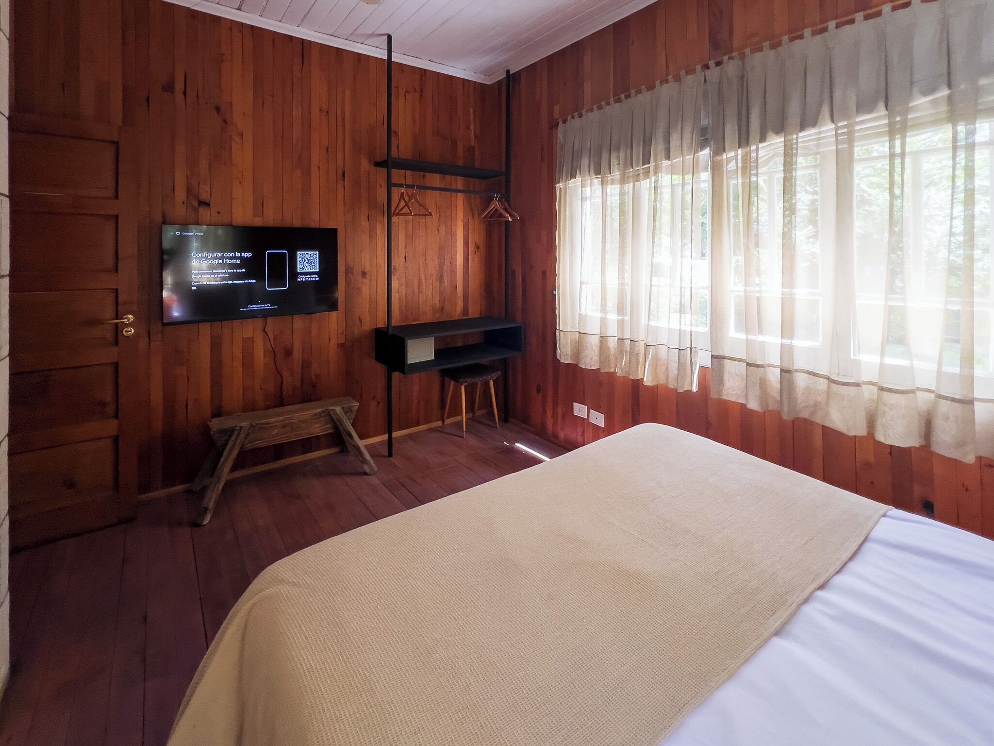 A wooden room with a bed, a TV mounted on the wall, a bench, and a window with curtains.