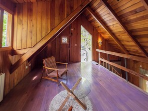 Interior de madeira com teto inclinado, uma cadeira, uma mesa pequena e vista através de uma porta.