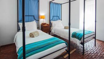 Una habitación con dos camas con dosel, cada una con una mesita de noche de madera y una lámpara. El suelo es de ladrillo y las paredes son blancas.