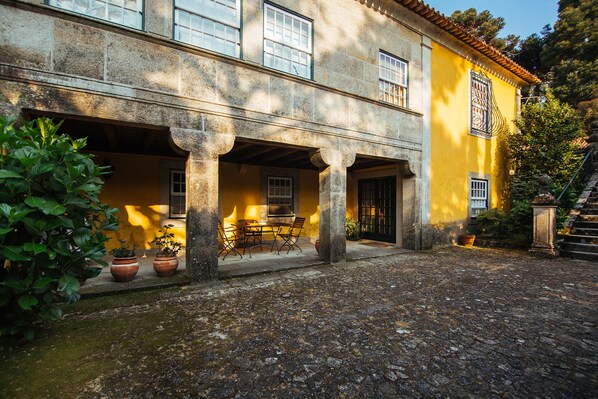 A stone building with a yellow facade, a covered patio with a table and chairs, and potted plants. - Quinta da Bouça d'Arques (Viana do Castelo)