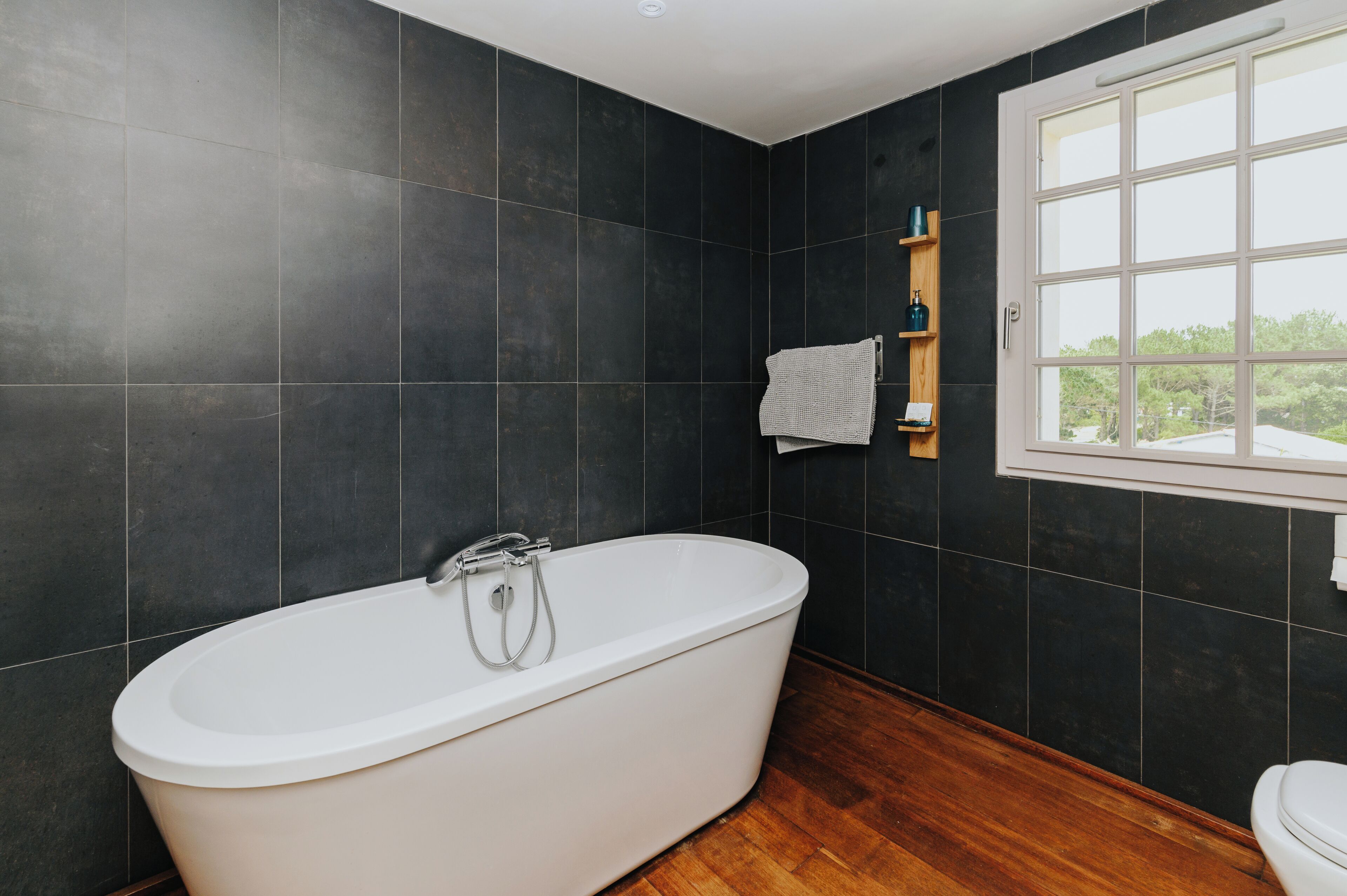 Une salle de bain moderne avec des carreaux foncés, une baignoire sur pieds, une étagère en bois où sont rangés les produits de toilette et une fenêtre offrant une vue agréable.