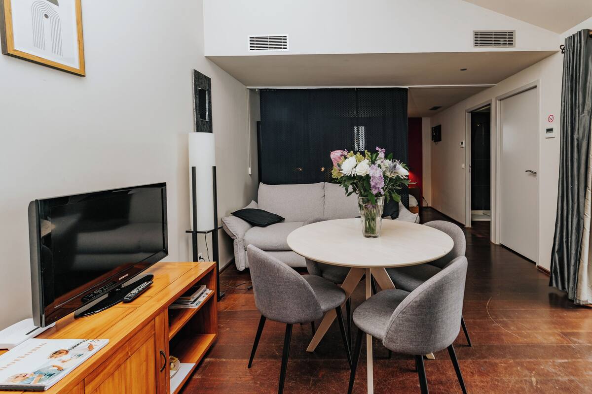 Une chambre d’hôtel moderne équipée d’un téléviseur à écran plat, d’un canapé, d’une table de salle à manger ronde avec des chaises et d’un vase de fleurs.