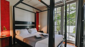 A four-poster bed with white linens, a bedside table with a lamp, and a view of greenery through large windows.